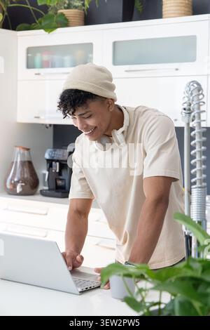 Afroamerikaner in 20ern, der sich in der Küche über einen Laptop beugt und mit Beanie und Kopfhörern lächelt Stockfoto
