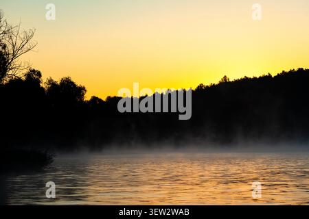 Die goldene Morgendämmerung leuchtet über einem ruhigen Waldsee mit sanftem Nebel, der über die Wellen strömt. Dunkle Baumsilhouetten säumen das Ufer, während sich warmes Licht über t ausbreitet Stockfoto