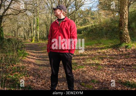 Mann mit roter Jacke und Mütze, der auf einem Waldweg steht, der mit Herbstlaub bedeckt ist Stockfoto