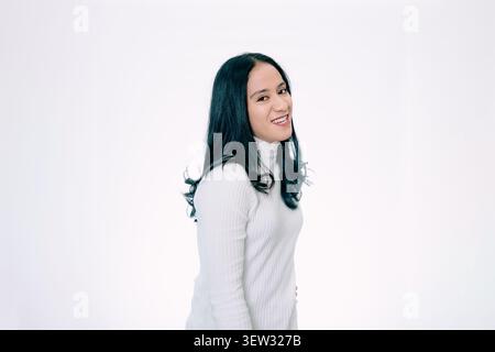 Lächelnde asiatische Frau, die die Kamera über ihrer Schulter auf weißem Hintergrund ansieht Stockfoto