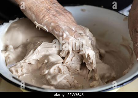 Ein reifer Einzelteil knettet klebrige Sauerteigblätter in einer Schüssel aus weißer Emaille und zeigt die haptische Zubereitung von organischen Zutaten in einem warmen tr Stockfoto