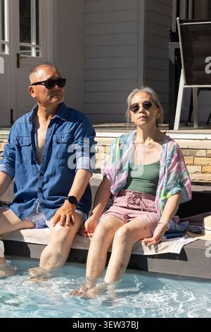 Verschiedene Paare sitzen auf dem Poolvorsprung im Hinterhof, Füße im Wasser, tragen Sonnenbrille und Denim Stockfoto