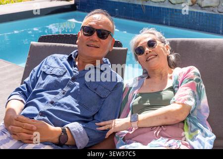 Asiatisches Seniorenpaar, das sich nebeneinander auf Liegestühlen am Hinterhof-Pool liegend und eine Sonnenbrille trug Stockfoto