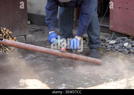 Eine Person, die blaue Schutzhandschuhe trägt, bedient einen elektrischen Winkelschleifer an einem Stahlrohr, um Korrosion zu entfernen, was wichtige Baureparaturen veranschaulicht Stockfoto