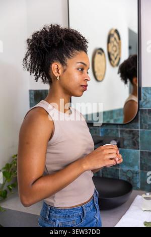 Afroamerikanerin hält ein Glas am Waschtisch im Badezimmer in beigefarbenem Tanktop und Jeans, Spiegel sichtbar Stockfoto