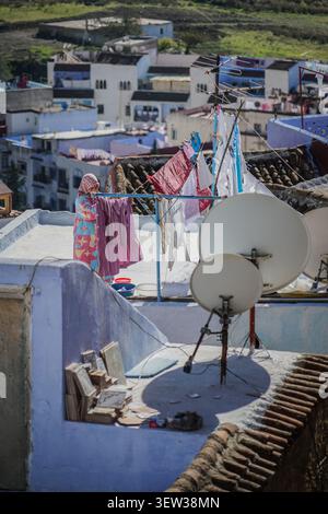 Chefchaouen, Marokko - 6. Dezember 2017: Eine Frau in einem farbenfrohen gemusterten Gewand und Kopftuch hängt lila Wäsche an einer Wäscheleine auf einem hellen Dach. Stockfoto