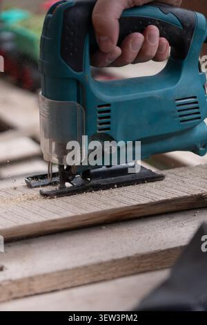 Eine detaillierte Nahaufnahme der Hände mit einer elektrischen Handstichsäge zum Schneiden einer rauen Holzdiele während einer Zimmerei oder einer Renovierung eines Heimwerkerwohnheims; Stockfoto