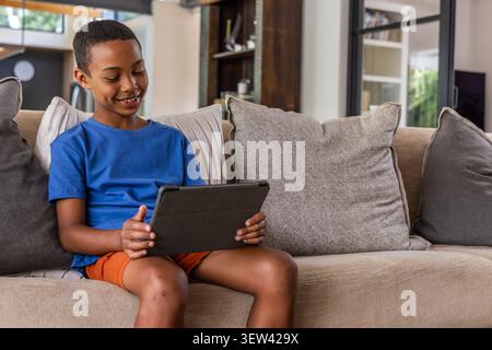 Afroamerikanischer Junge in blauem Hemd, orangen Shorts, der auf dem Sofa sitzt und eine Tablette in der Schutzhülle hält Stockfoto