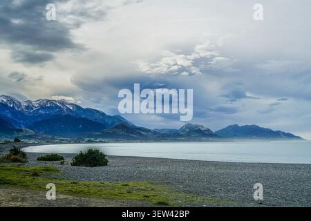 Eine breite Landschaftsansicht der dramatischen Küste von Kaikoura, mit einem ruhigen Pazifik, einem Kiesstrand im Vordergrund und der majestätischen schneebedeckten majestätischen Landschaft Stockfoto