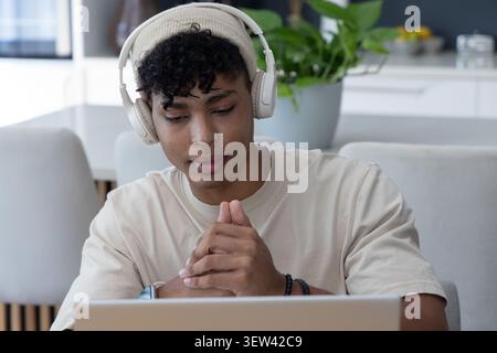 Afroamerikaner in den Zwanzigern mit beigefarbener Mütze und Kopfhörern, der in der Küche an einem Laptop arbeitet Stockfoto