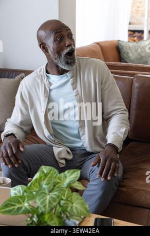 Ranghoher Afroamerikaner sitzt zu Hause auf Ledercouch, trägt beiges Hemd und hält Smartphone Stockfoto
