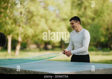 Ein Mann, der ein Tischtennisnetz auf einem Betontisch im Freien in einem sonnigen Park aufstellt Stockfoto