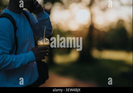 Junger Mann mit Rucksack, der telefoniert und einen Drink in einem Park bei Sonnenuntergang hält Stockfoto
