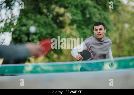 Junger Mann, der draußen Tischtennis spielt, mit seinem Gegner an einem Tischtennistisch im Park Stockfoto