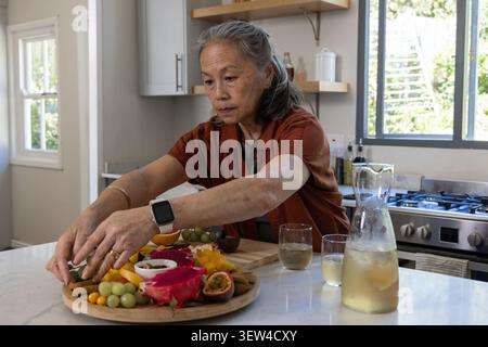 Ältere asiatische Frau im Rosthemd, die auf der Marmorinsel eine Platte mit Drachenfrüchten und Trauben arrangiert Stockfoto