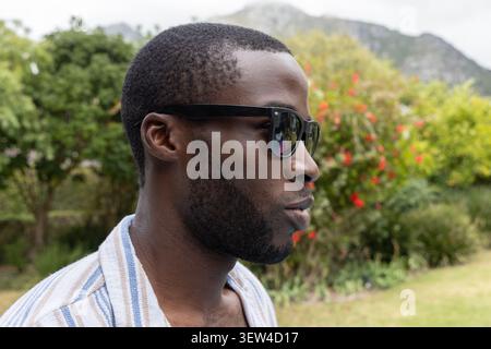 Afroamerikaner, der Sonnenbrille trägt, gestreiftes Hemd, steht im Park mit roten Blüten Stockfoto