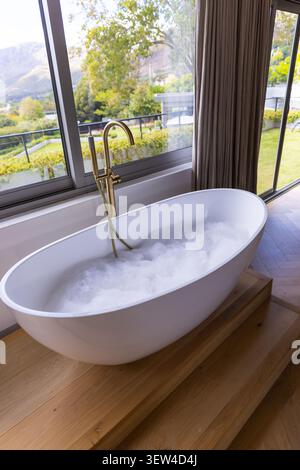 Die ovale weiße Badewanne befindet sich auf der Plattform im Badezimmer in der Nähe von Fenstern mit Messingarmatur und Schaum Stockfoto