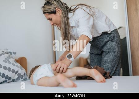 Die Mutter wechselt die Windel auf einem Kleinkind auf dem Bett in einem hellen Schlafzimmer, sorgenvoller Elternmoment Stockfoto