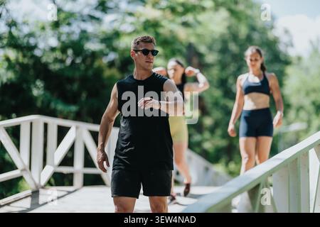 Ein Mann führt einen Outdoor-Fitness-Spaziergang auf einer sonnigen Brücke mit zwei Frauen, die in Sportbekleidung nachgehen Stockfoto