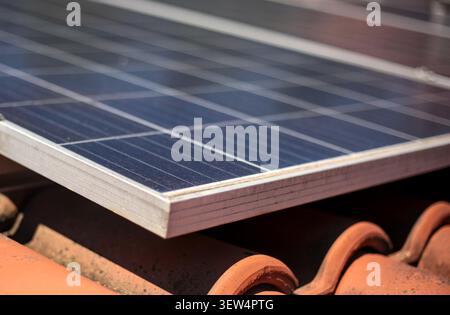 Eine fokussierte Nahaufnahme, die die metallischen Ecken und Kanten eines modernen Solarmoduls zeigt, der direkt auf verwitterten roten Lehm-Dachziegeln montiert ist. Stockfoto