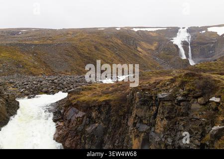 Malerischer Blick auf den Wasserfall Rjúkandi, der abgelegene felsige Hügel und Klippen hinunter in einen rauschenden Fluss unter einem bewölkten Himmel in Island stürzt. Stockfoto