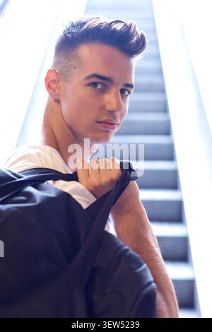 Kaukasischer junger Mann in schwarzem Hemd posiert mit Händen in den Haaren neben alten Fenstern, blickt nach oben im warmen Sonnenlicht, Mode-Lifestyle-Porträt. Stockfoto