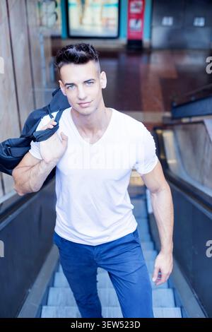 Kaukasischer junger Mann in weißem Hemd und blauer Hose fährt drinnen auf einer Rolltreppe, trägt einen Rucksack und schaut in die Kamera. Stockfoto