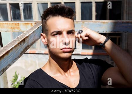 Kaukasischer junger Mann in schwarzem Hemd posiert draußen neben einem rostigen Geländer, trägt Schmuck und sieht im direkten Sonnenlicht ernst aus. Stockfoto