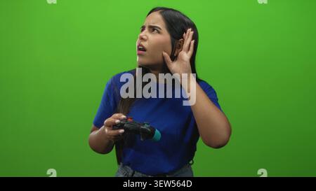 Frau, die mit der rechten Hand Gamepad hält, während sie im Studio von der linken Hand an das Ohr schwingt, zuhört und zuhört, als würde sie Audiosignale hören; Verwirrtheit Neugier. Stockfoto