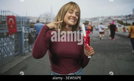 Frau mit Diplom zeigt Finger an Wange auf der Straße, lächelt mit sichtbaren Zähnen und rotem Band auf der Schriftrolle; Stolz auf Leistung. Stockfoto