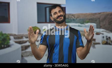 Mann in blau gestreifter Fußballuniform mit Bart hebt offene Hände, Palmen sichtbar, vor dem Gebäude; amüsierte Leichtigkeit. Stockfoto