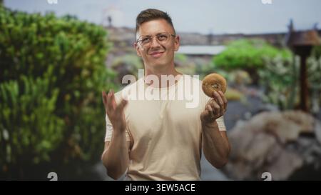 Ein Mann hält einen Bagel mit einer Hand und ballt die Faust im Studio, zuzwinkernd und lächelnd mit geschlossenen Augen, als ob er einen kräftigen Biss schmecken würde; Vergnügungssucht. Stockfoto