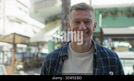 Mann in einem blauen karierten Hemd lächelt in die Kamera neben einem weißen Arbeitswagen mit Dachträger auf einer sonnendurchfluteten Fußgängerzone, die mit Cafés und Sonnenschirmen gesäumt ist; warm Stockfoto