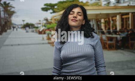 Junge brünette Frau, die mit geschlossenen Augen und einem leichten Lächeln auf der Straße neben einer Restaurantterrasse im Freien steht; ruhige Reflexion. Stockfoto