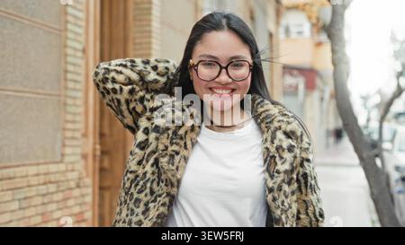 Frau mit Brille und Leopardenpelz lächelt mit der Hand hinter dem Kopf auf der Straße und lehnt sich an ein Gebäude; ungezwungenes Vertrauen. Stockfoto