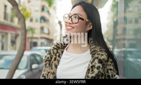 Frau mit Schildpatt-Brille und Leopardenmantel lächelt und blickt auf eine Stadtstraße in der Nähe geparkter Autos und Glasfassaden; Optimismus. Stockfoto