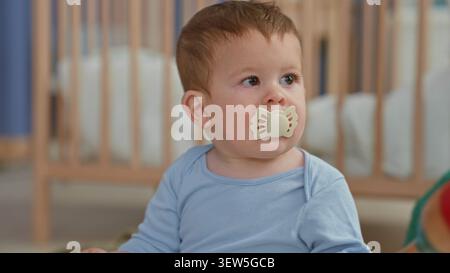 Kleinkind mit Schnuller, der rote Rassel hält und mit der Hand greift, während er in der Nähe einer Holzkrippe im Kinderzimmer sitzt; ruhige Neugier. Stockfoto