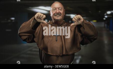 Mönch in brauner Angewohnheit mit Kreuz-Halskette zeigt beide Daumen nach unten mit sichtbaren Händen und ein Lächeln in einem dunklen Tiefgaragengebäude Stockfoto