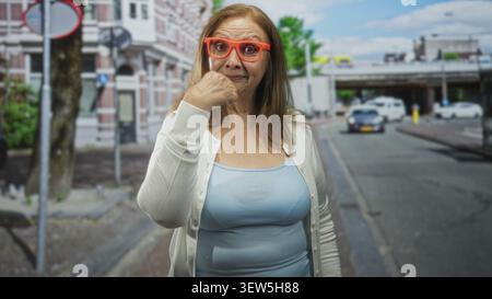 Frau mit orangefarbener Brille, die auf einer Straße vor Gebäuden ihren Mund berührt, in Strickjacke und Tanktop; nachdenkliche Reflexion. Stockfoto