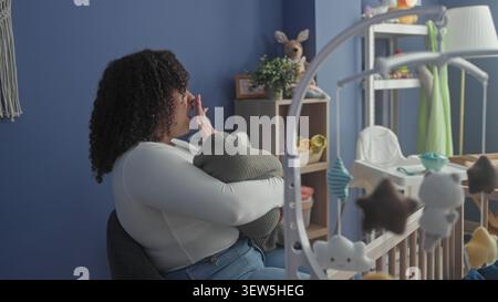 Die Frau wiegt das Baby in eine graue Decke, reibt die Augen und legt den Kopf neben dem Kinderbett mit einem gefüllten Häschen an die blaue Wand im Gebäude; Erschöpfungströst. Stockfoto