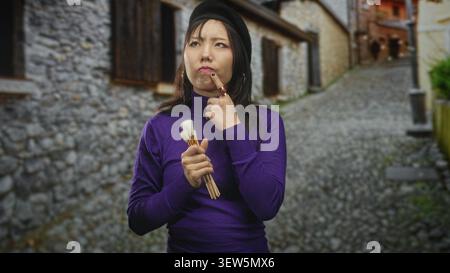 Junge chinesin, die Pinsel hält, Finger bis Kinn, Baskenmütze und lila Pullover auf der Kopfsteinpflasterstraße trägt; leises Denken. Stockfoto
