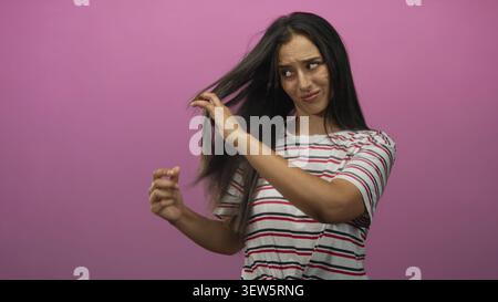 Frau, die langes Haar hält und untersucht, auf Spliss im Studio mit rosafarbenem Hintergrund; Haarpflege-Frustration. Stockfoto