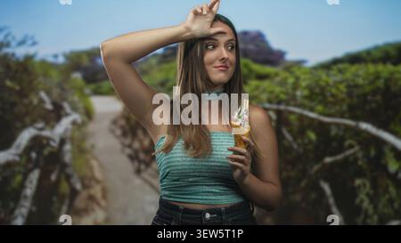 Die junge brünette Frau, die Eisbecher hält, lässt mich auf der Stirn ein Zeichen setzen, während sie auf einem Waldweg in der Nähe eines hölzernen Geländes steht; verspielter Sommerspaß. Stockfoto