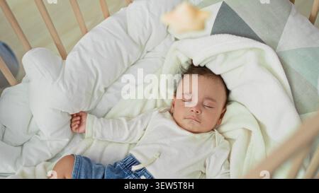 Hispanischer Kleinkind, der auf dem Rücken in einem hölzernen Wiegenbett mit weißer Decke und Jeansshorts schläft, Hand auf Kissen im Kinderzimmer gerollt; friedliche Ruhe. Stockfoto