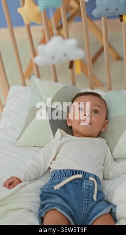 Hispanischer Junge, der im Kinderbett auf dem Bett liegt, Kopf auf gemustertem Kissen, trägt Jeansshorts und blickt auf das Plüschmobil, das über Holzlatten hängt Stockfoto
