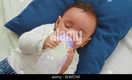 Baby Boy hält Milchflasche und stillt, während er auf dem Bett im Gebäude weint; verstörte beruhigende Komfortpflege. Stockfoto