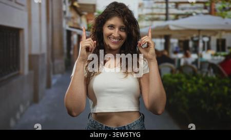 Die Frau zeigt beide Zeigefinger auf eine Straßenterrasse, lächelt mit sichtbarem Mittelriff; Freude. Stockfoto