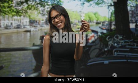 Thailändische Frau hält ein weißes Puzzleteil mit der Hand nahe ihrem Gesicht auf einer Straße an einem Kanal mit Fahrrädern und Bäumen; Neugier. Stockfoto