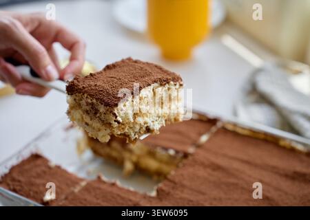 Hebt die Tiramisu-Scheibe mit einem Spachtel von der Hand an, der mit Kakaopulver bedeckt ist. Konzept des hausgemachten Desserts und Servierens Stockfoto