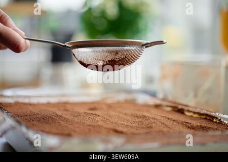 Die Hand hält das Sieb über dem mit Kakaopulver bedeckten Tiramisu-Dessert in einer Glasschale. Konzept des hausgemachten Desserts Stockfoto